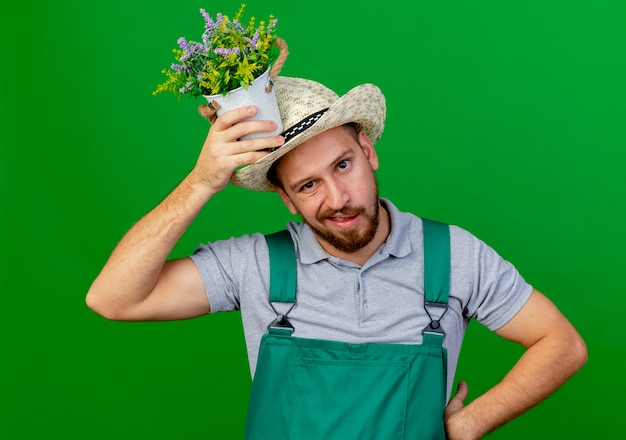 Insatisfait Jeune Beau Jardinier Slave En Uniforme Et Hat Holding Pot De Fleurs Sur La Tête En Gardant La Main Sur La Taille à La Recherche