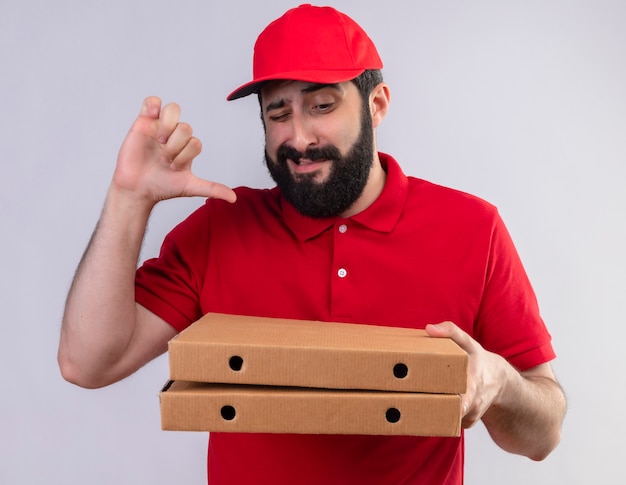 Insatisfait jeune beau livreur caucasien vêtu d'un uniforme rouge et cap tenant et regardant des boîtes de pizza et montrant le pouce vers le bas isolé sur fond blanc