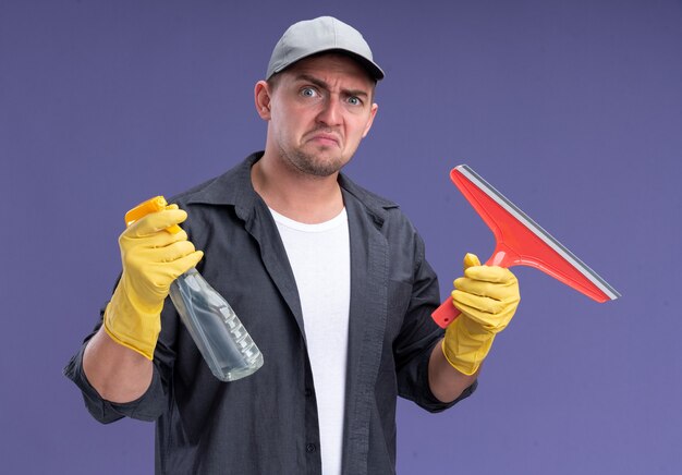 Insatisfait jeune beau mec de nettoyage portant t-shirt avec capuchon et gants tenant la tête de vadrouille et vaporisateur isolé sur mur violet