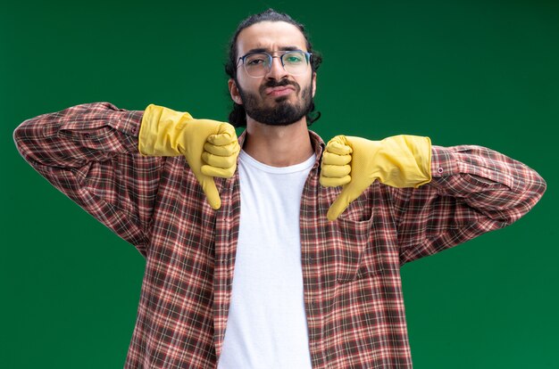 Insatisfait jeune beau mec de nettoyage portant un t-shirt et des gants montrant les pouces vers le bas isolé sur un mur vert