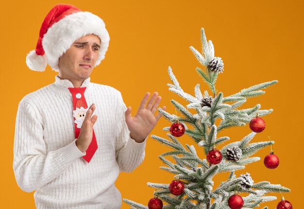 Insatisfait jeune beau mec portant chapeau de Noël et cravate de père Noël debout près de sapin de Noël décoré en le regardant faire le geste de refus isolé sur fond orange