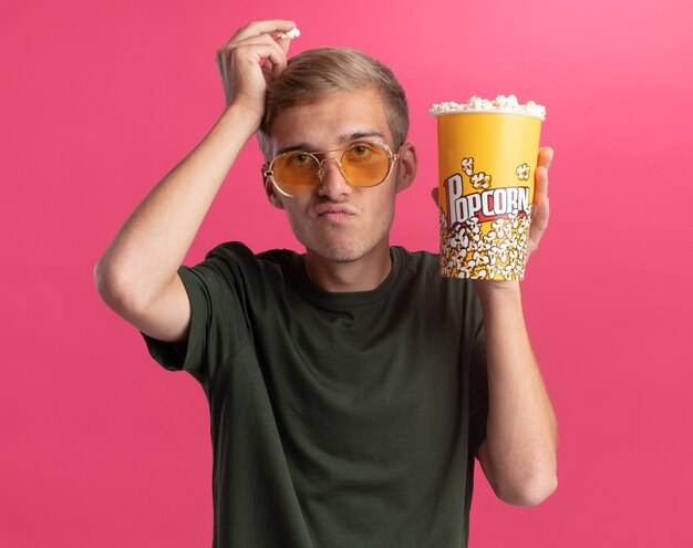 Insatisfait jeune beau mec portant une chemise verte et des lunettes tenant un seau de pop-corn et tenant un morceau de pop-corn sur la tête isolé sur un mur rose