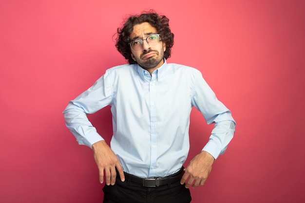 Insatisfait jeune bel homme portant des lunettes à l'avant en gardant les mains en l'air isolé sur un mur rose