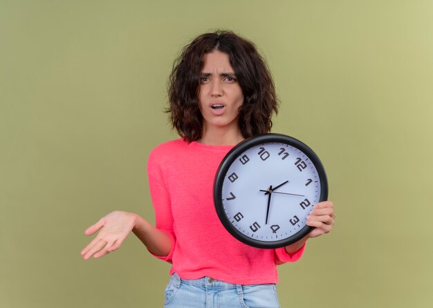 Insatisfait jeune belle fille tenant horloge et montrant la main vide sur fond vert isolé