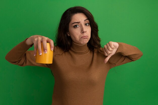 Insatisfait jeune belle fille tenant une tasse de thé et montrant le pouce vers le bas isolé sur un mur vert