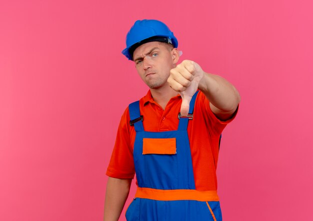 Insatisfait jeune constructeur masculin portant l'uniforme et un casque de sécurité son pouce vers le bas sur rose