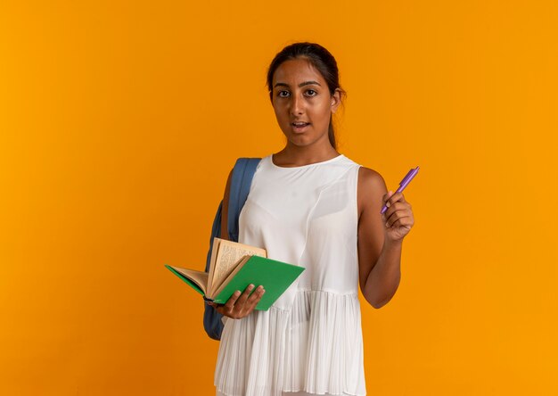 Insatisfait jeune écolière portant un sac à dos tenant un livre et un stylo sur orange