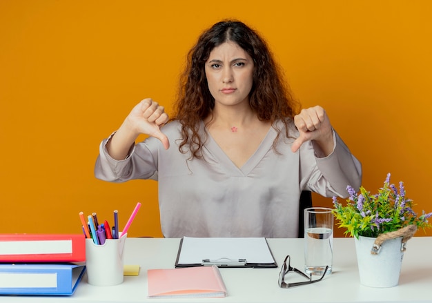 Insatisfait jeune employé de bureau jolie femme assis au bureau avec des outils de bureau ses pouces vers le bas isolé sur orange