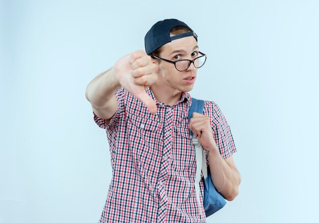 Insatisfait jeune étudiant garçon portant un sac à dos et des lunettes et casquette son pouce vers le bas sur blanc