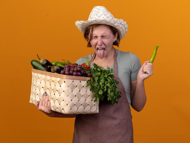 Insatisfait jeune femme jardinier slave portant chapeau de jardinage sort la langue tenant le panier de légumes et le piment sur orange