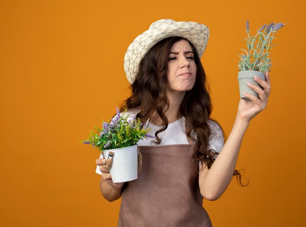 Insatisfait jeune femme jardinière en uniforme portant chapeau de jardinage détient des pots de fleurs isolés sur un mur orange avec copie espace