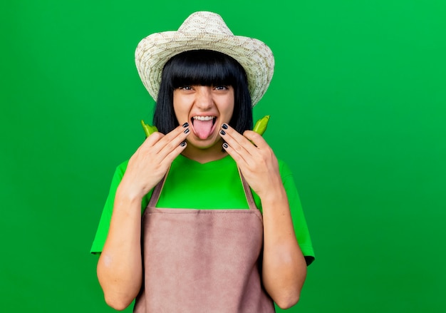 Insatisfait jeune femme jardinière en uniforme portant chapeau de jardinage sort la langue tient le piment cassé