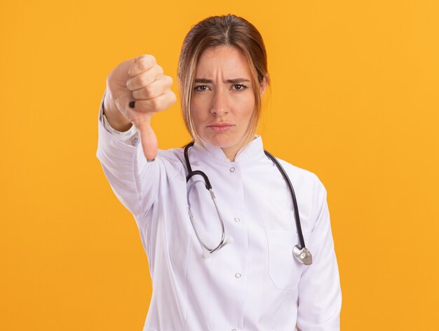 Insatisfait jeune femme médecin portant une robe médicale avec stéthoscope montrant le pouce vers le bas isolé sur mur jaune