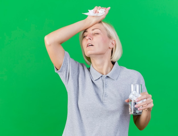 Insatisfait jeune femme slave malade blonde met la main sur le front détient un verre d'eau et pack de pilules médicales isolé sur un mur vert avec espace copie