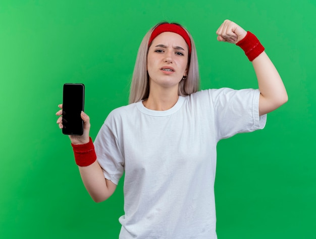 Insatisfait jeune femme sportive avec des accolades portant un bandeau et des bracelets se tient avec le poing levé et tient le téléphone isolé sur le mur vert