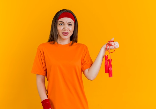 Insatisfait jeune femme sportive portant un bandeau et des bracelets tenant une corde à sauter isolée sur un mur orange avec espace de copie