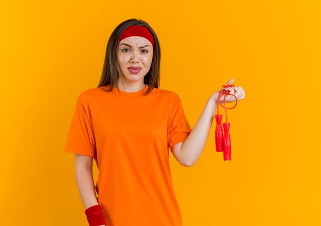 Insatisfait jeune femme sportive portant un bandeau et des bracelets tenant une corde à sauter isolée sur un mur orange avec espace de copie