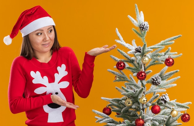 Insatisfait jeune fille asiatique portant chapeau de Noël avec chandail debout à proximité de l'arbre de Noël isolé sur mur orange