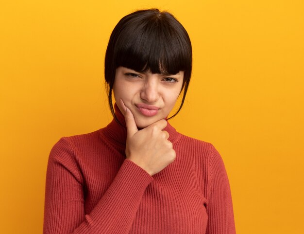 Insatisfait jeune fille caucasienne brune met la main sur le menton et regarde la caméra sur l'orange