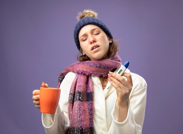 Insatisfait jeune fille malade aux yeux fermés portant une robe blanche et un chapeau d'hiver avec écharpe tenant une tasse de thé et un thermomètre isolé sur violet