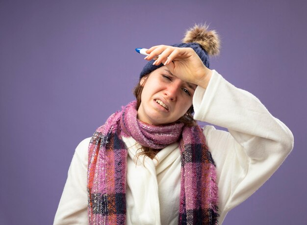 Insatisfait jeune fille malade portant une robe blanche et un chapeau d'hiver avec écharpe tenant un thermomètre mettant le bras sur le front isolé sur violet