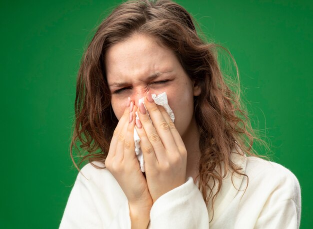Insatisfait jeune fille malade portant une robe blanche essuyant le nez avec serviette isolé sur vert