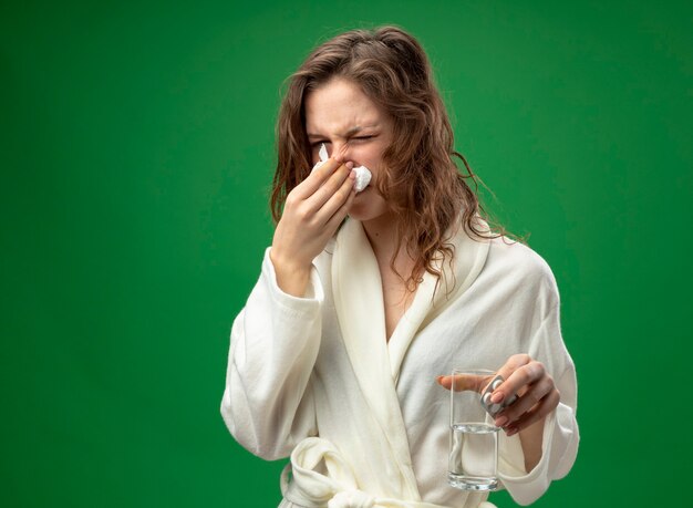 Insatisfait jeune fille malade portant une robe blanche tenant un verre d'eau en essuyant le nez avec une serviette isolé sur vert