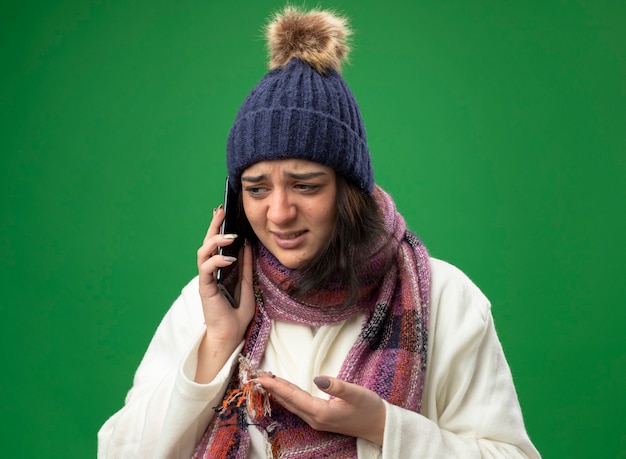 Insatisfait Jeune Fille Malade De Race Blanche Portant Chapeau D'hiver Robe Et écharpe Parler Au Téléphone Montrant La Main Vide à Côté Isolé Sur Mur Vert