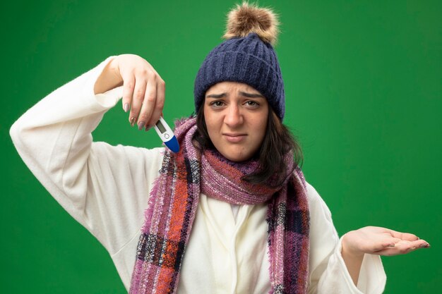 Insatisfait jeune fille malade de race blanche portant chapeau d'hiver robe et écharpe tenant un thermomètre montrant la main vide regardant la caméra isolée sur fond vert