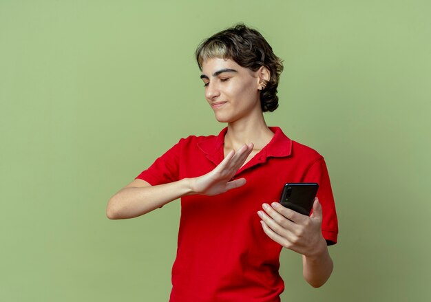 Insatisfait jeune fille de race blanche avec coupe de cheveux de lutin tenant le téléphone mobile et faisant des gestes non avec les yeux fermés isolé sur fond vert olive avec espace copie