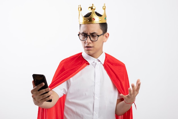 Insatisfait jeune garçon de super-héros en cape rouge portant des lunettes et une couronne tenant et regardant le téléphone mobile en gardant la main dans l'air isolé sur fond blanc