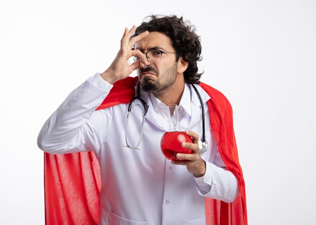 Insatisfait jeune homme caucasien dans des lunettes optiques portant l'uniforme du médecin avec manteau rouge et avec stéthoscope autour du cou ferme le nez et détient un liquide chimique rouge dans un flacon de verre sur un mur blanc