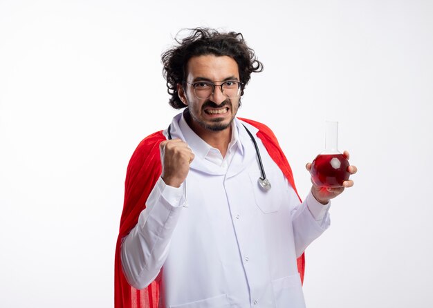 Insatisfait jeune homme caucasien dans des lunettes optiques portant l'uniforme de médecin avec manteau rouge et avec stéthoscope autour du cou garde le poing et détient un liquide chimique rouge dans un flacon de verre sur un mur blanc