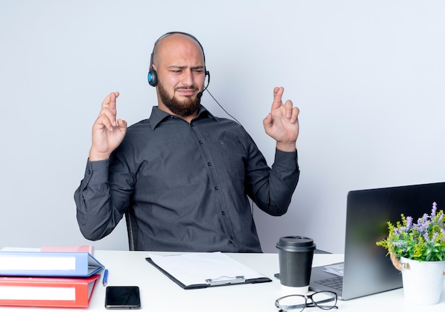 Insatisfait jeune homme de centre d'appels chauve portant un casque assis au bureau avec des outils de travail à l'ordinateur portable avec les doigts croisés isolé sur fond blanc