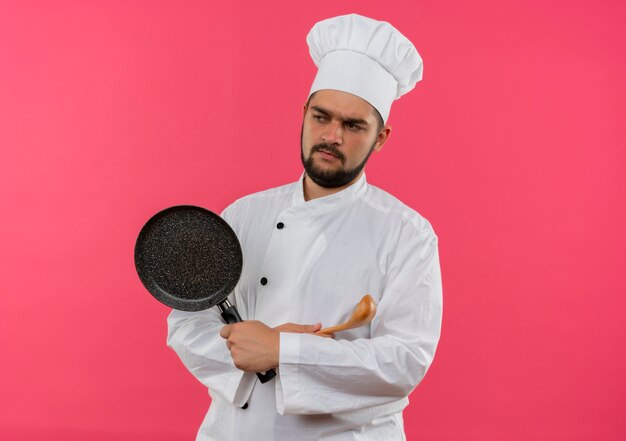 Insatisfait jeune homme cuisinier en uniforme de chef debout avec une posture fermée et tenant une poêle avec une cuillère à côté isolé sur l'espace rose