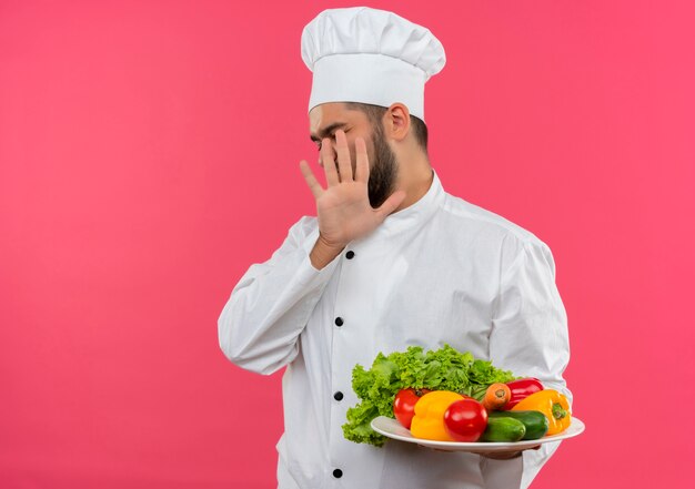 Insatisfait jeune homme cuisinier en uniforme de chef tenant une assiette de légumes et faisant des gestes pas isolé sur l'espace rose