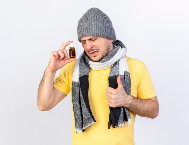 Insatisfait jeune homme malade blonde portant un chapeau d'hiver et une écharpe détient des médicaments dans une bouteille en verre et les pouces vers le haut isolé sur un mur blanc