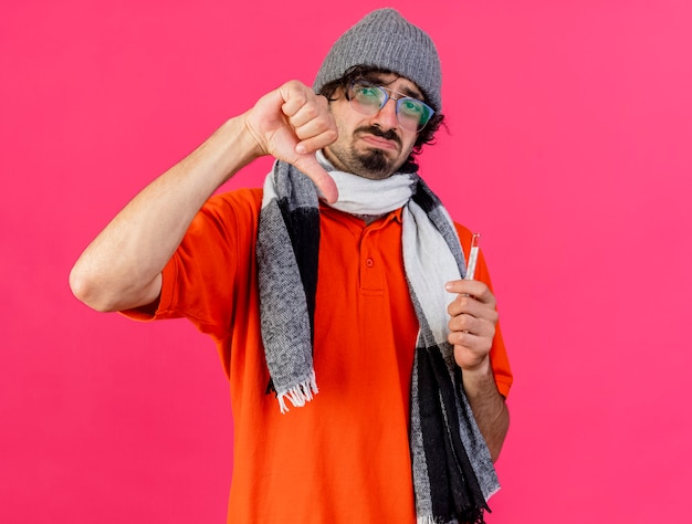 Photo gratuite insatisfait jeune homme malade de race blanche portant des lunettes chapeau d'hiver et écharpe tenant un thermomètre regardant la caméra montrant le pouce vers le bas isolé sur fond cramoisi avec espace de copie