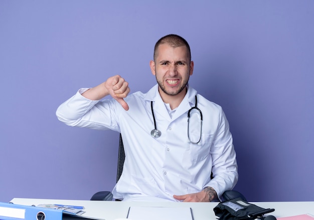 Insatisfait jeune homme médecin portant robe médicale et stéthoscope assis au bureau avec des outils de travail montrant le pouce vers le bas isolé sur fond violet