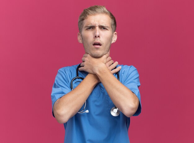 Insatisfait jeune homme médecin portant l'uniforme de médecin avec stéthoscope a attrapé la gorge isolé sur mur rose