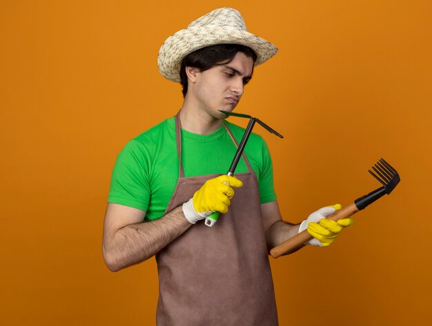 Insatisfait jeune jardinier mâle en uniforme portant chapeau de jardinage avec des gants tenant et regardant houe râteau avec râteau isolé sur orange