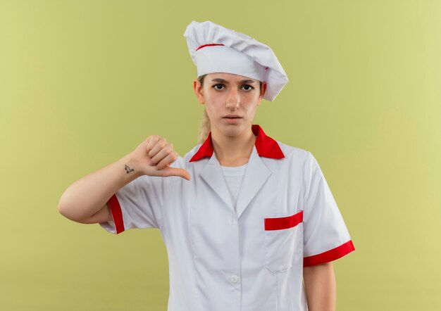 Insatisfait jeune jolie cuisinier en uniforme de chef montrant le pouce vers le bas à isolé sur l'espace vert