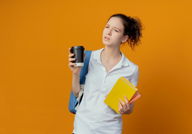 Insatisfait jeune jolie étudiante portant sac à dos tenant le stylo bloc-notes livre et tasse à café en plastique à côté isolé sur fond orange avec copie espace