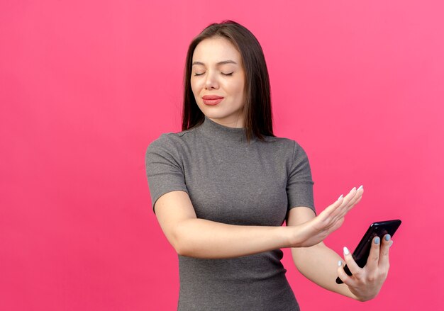 Insatisfait jeune jolie femme tenant un téléphone mobile et faisant des gestes non avec les yeux fermés isolé sur fond rose avec espace de copie