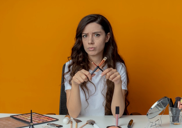 Insatisfait jeune jolie fille assise à la table de maquillage avec des outils de maquillage tenant des pinceaux de maquillage et faisant des gestes non avec eux à la caméra isolée sur fond orange