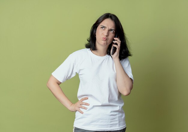 Insatisfait jeune jolie fille caucasienne parlant au téléphone en regardant côté mettre la main sur la taille isolé sur fond vert olive avec espace copie