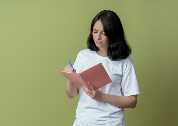 Insatisfait jeune jolie fille caucasienne tenant un stylo et un bloc-notes écrit quelque chose d'isolé sur fond vert olive avec espace de copie