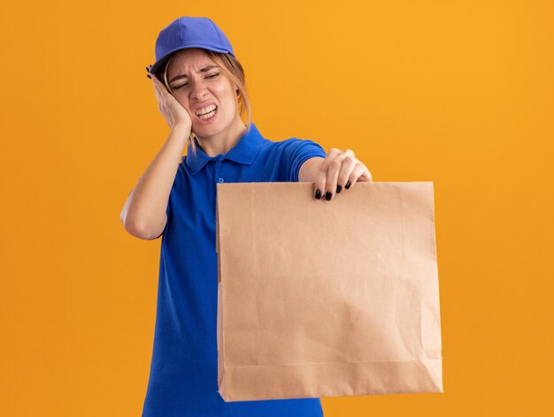 Insatisfait jeune jolie livreuse en uniforme met la main sur le visage et détient un paquet de papier sur orange