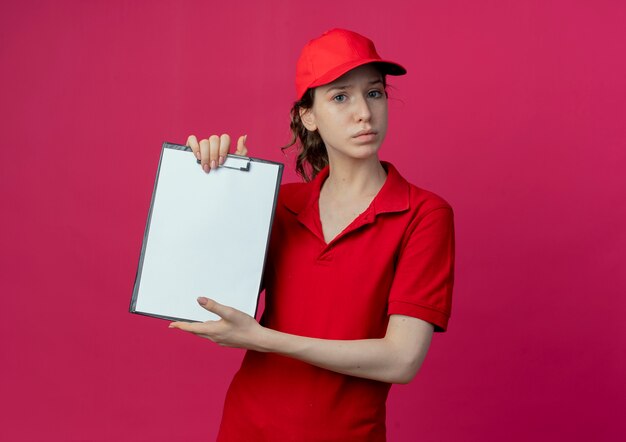 Insatisfait jeune jolie livreuse en uniforme rouge et cap montrant le presse-papiers isolé sur fond cramoisi avec espace de copie