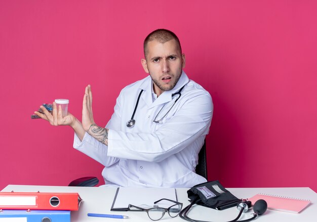 Photo gratuite insatisfait jeune médecin de sexe masculin portant une robe médicale et un stéthoscope assis au bureau avec des outils de travail tenant un bécher médical et des paquets de comprimés et de capsules et de gestes non isolé sur fond rose